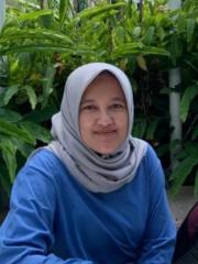 A headshot of Fitrah – an Indonesian woman with an oval face and warm brown skin, wears a headscarf and is sitting in front of green plants