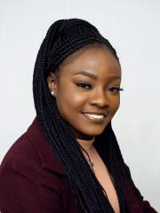A headshot of Pelumi Oginni - A brown-skinned woman with long black braided hair wearing a burgundy blazer over a peach-coloured top