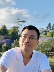 Headshot of Minh, a man with short dark hair and glasses, wearing a white shirt and smiling slightly, set against a lush outdoor background with trees and a blue sky