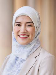 •	A headshot of Nena - a Southeast Asian women with head covering/hijab smiling and standing in front of a blurred building background