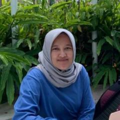 A headshot of Fitrah – an Indonesian woman with an oval face and warm brown skin, wears a headscarf and is sitting in front of green plants