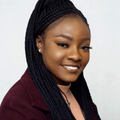A headshot of Pelumi Oginni - A brown-skinned woman with long black braided hair wearing a burgundy blazer over a peach-coloured top