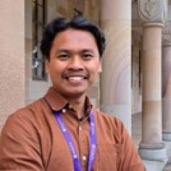 A headshot of Juddi – a smiling Javanese Indonesian man with short, dark-black, wavy hair standing at UQ's Great Court