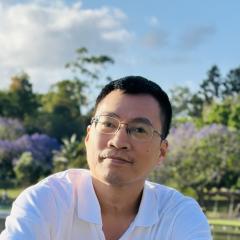 Headshot of Minh, a man with short dark hair and glasses, wearing a white shirt and smiling slightly, set against a lush outdoor background with trees and a blue sky