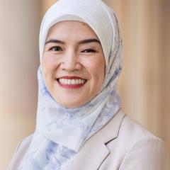•	A headshot of Nena - a Southeast Asian women with head covering/hijab smiling and standing in front of a blurred building background